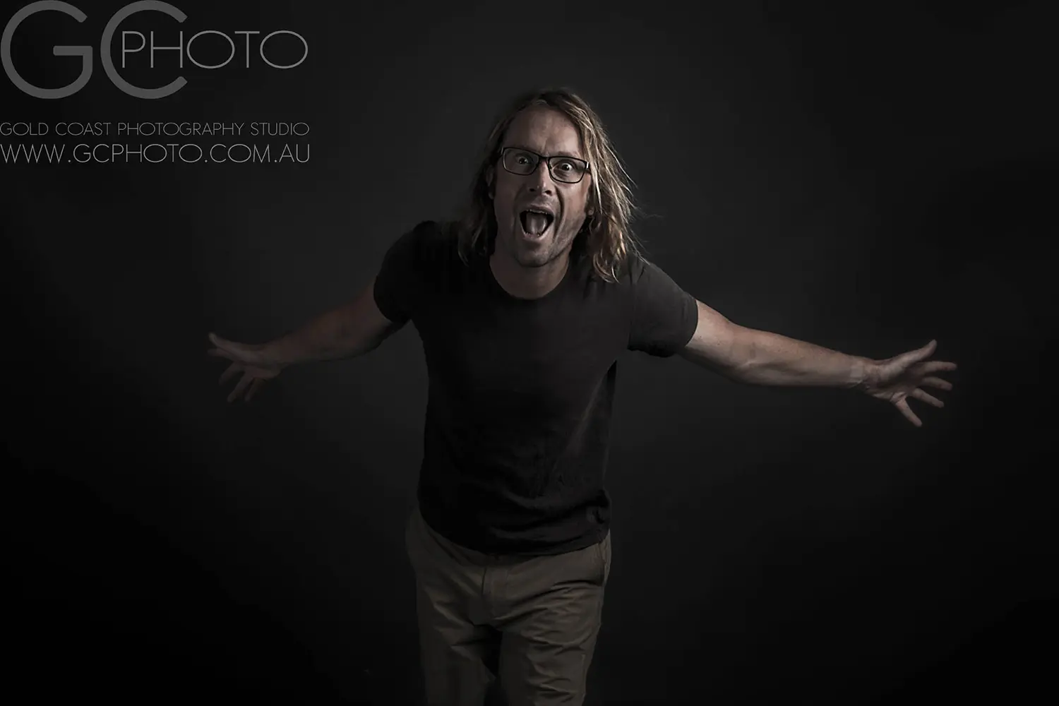 Long-haired man in glasses spreading arms against dark background